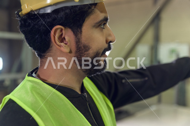 عامل مسؤول ومشرف سعودي يرتدي خوذه صفراء يشرف داخل المصنع في المملكة العربية السعودية , ارتداء خوذة وسترة الحماية الخاصة بالعمل , التصنيع والإشراف والتدقيق في العمل , الهندسة والصناعة
