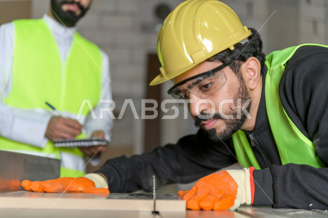 Two Saudi engineers working supervising inside the factory in Saudi Arabia, wearing a work helmet and protective jacket, the responsible worker in a yellow helmet, the supervising engineer in a white helmet, checking the equipment themselves, checking the