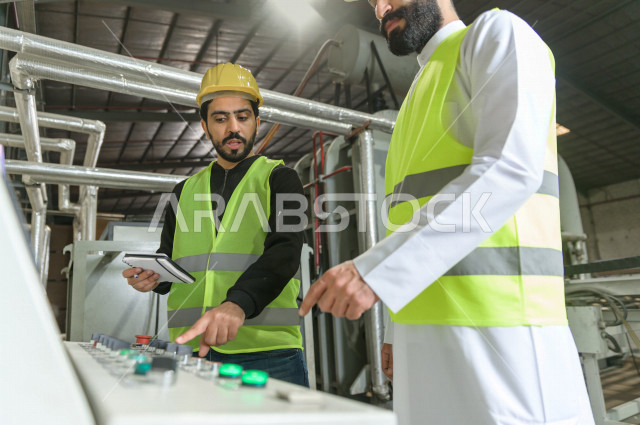 Two Saudi engineers working supervising inside the factory in Saudi Arabia, wearing a work helmet and protective jacket, the responsible worker in a yellow helmet, the supervising engineer in a white helmet, checking the equipment themselves, checking the