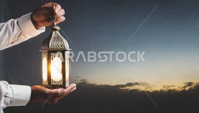 Lighting and decorations for the holy month of Ramadan, lanterns and Islamic artifacts to celebrate the arrival of the month of goodness, a close-up of the hands of a Saudi Arabian Gulf man holding a lit metal lantern, decorations and lights in the spiritual Ramadan atmosphere
