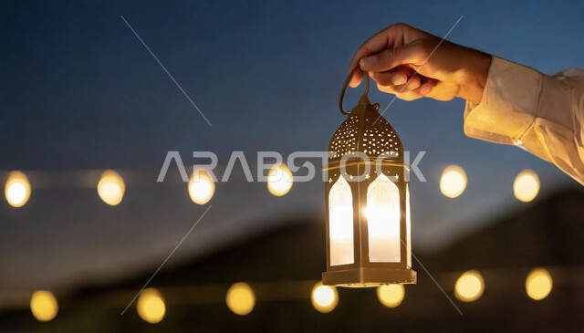  ديكورات وأنوار في الأجواء الرمضانية الروحانية، إضاءة وزينة شهر رمضان المبارك، فوانيس وتحف اسلامية للاحتفال بقدوم شهر الخير، صورة مقربة لأيدي رجل عربي خليجي سعودي يحمل فانوس معدني مضيء