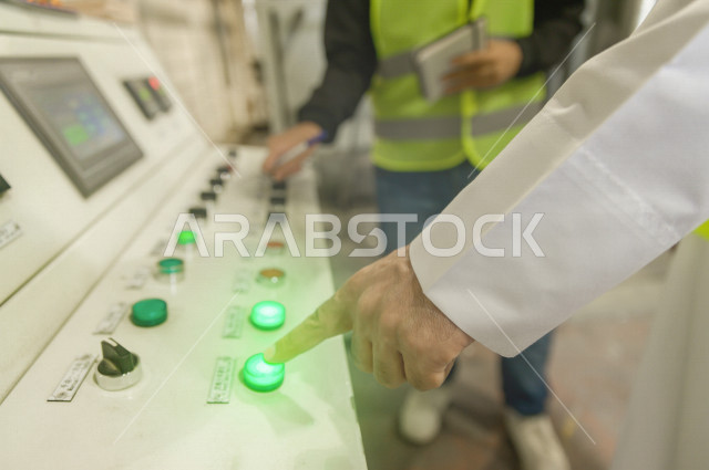 صورة مقربة ليد المهندسان السعوديان العاملان يقومون بالإشراف في داخل المصنع في المملكة العربية السعودية , إرتداء سترة الحماية الخاصة بالعمل , فحص المعدات بأنفسهم , تدقيق العمل , الإشراف والنقاش في العمل , لوحة أزرار التحكم , كيفية آلية التشغيل في المصنع