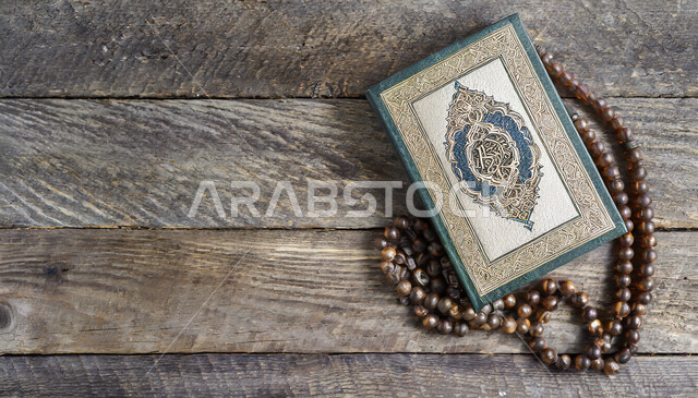 صورة علوية عامودية للمصحف الشريف بجانب سبحه مصنوعه من الخرز لذكر الله تعالى على لوح خشبي, التعبد والاجتهاد بالطاعة بتلاوة القران و ذكر الله, الهداية بنور القرآن الكريم, حفظ و تفسير آيات الكتاب الشريف