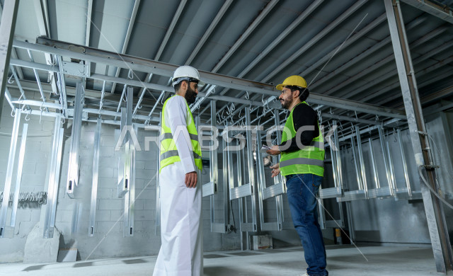 Two Saudi engineers working supervising inside the factory in Saudi Arabia, wearing a work helmet and protective jacket, the responsible worker in a yellow helmet, the supervising engineer in a white helmet, checking the equipment themselves, checking the