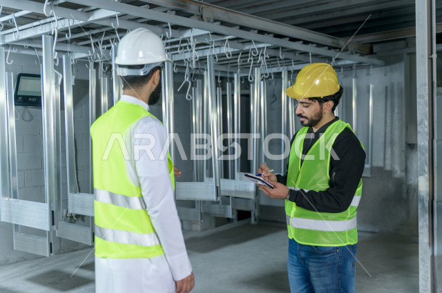 مهندسان  سعوديان عاملان يشرفان في داخل المصنع في المملكة العربية السعودية , إرتداء خوذة وسترة الحماية الخاصة بالعمل , العامل المسؤول بخوذة صفراء , المهندس المشرف بخوذة بيضاء , فحص المعدات بأنفسهم , تدقيق العمل , الإشراف والنقاش في العمل