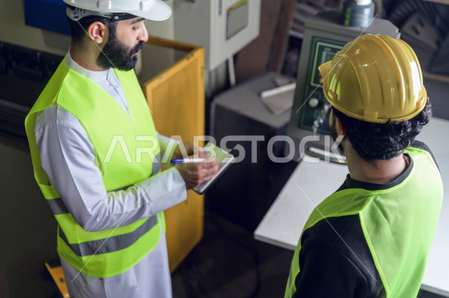 مهندسان  سعوديان عاملان يشرفان في داخل المصنع في المملكة العربية السعودية , إرتداء خوذة وسترة الحماية الخاصة بالعمل , العامل المسؤول بخوذة صفراء , المهندس المشرف بخوذة بيضاء , فحص المعدات بأنفسهم , تدقيق العمل , الإشراف والنقاش في العمل