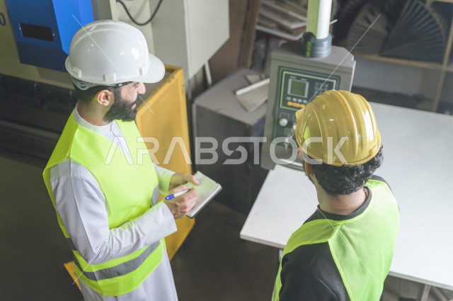 مهندسان  سعوديان عاملان يشرفان في داخل المصنع في المملكة العربية السعودية , إرتداء خوذة وسترة الحماية الخاصة بالعمل , العامل المسؤول بخوذة صفراء , المهندس المشرف بخوذة بيضاء , فحص المعدات بأنفسهم , تدقيق العمل , الإشراف والنقاش في العمل