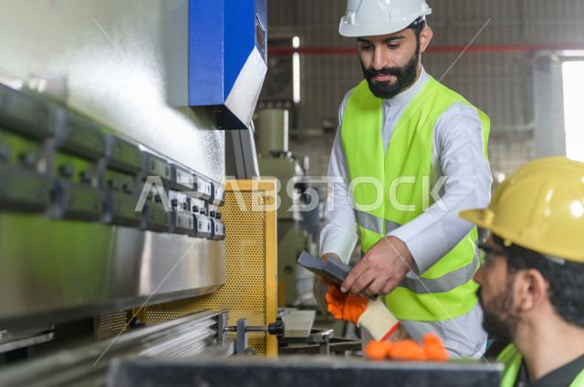 مهندسان  سعوديان عاملان يشرفان في داخل المصنع في المملكة العربية السعودية , إرتداء خوذة وسترة الحماية الخاصة بالعمل , العامل المسؤول بخوذة صفراء , المهندس المشرف بخوذة بيضاء , فحص المعدات بأنفسهم , تدقيق العمل , الإشراف والنقاش في العمل