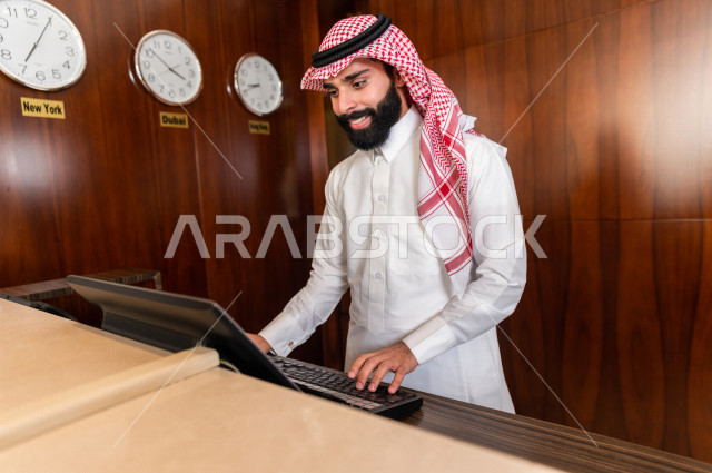 A smiling Saudi Gulf receptionist records data through the keyboard with different facial and hands gestures, an elegant man wearing a white dress with a shemagh agal, tourism and hotel, customer reception desk