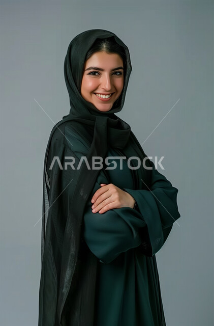 الملامح العربية القوية، أزياء محجبات عصرية وأنيقة، مفهوم الأنوثة والنعومة، الاهتمام بالمظهر الخارجي، بورتريه من الجانب لشابةعربية خليجية سعودية ترتدي العباية والحجاب تقف بأيدي متقاطعة، النظر الى الكاميرا بإيماءات البهجة والسرور، خلفية رمادية
