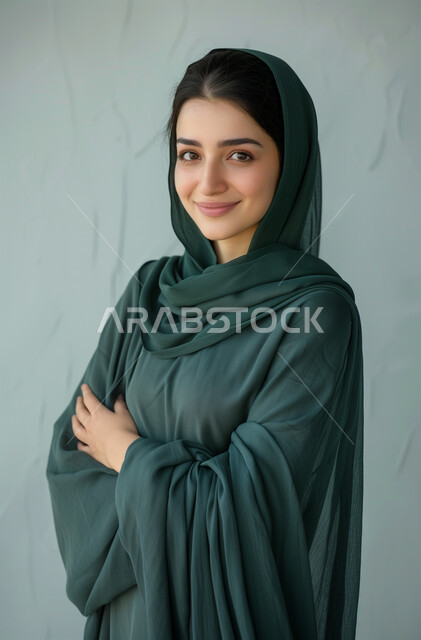 مفهوم الأنوثة والنعومة، الملامح العربية القوية، أزياء محجبات عصرية وأنيقة، الاهتمام بالمظهر الخارجي، بورتريه لشابة عربية خليجية سعودية ترتدي العباية والحجاب تقف بأيدي متقاطعة، النظر الى الكاميرا بإيماءات البهجة والسرور، خلفية رمادية