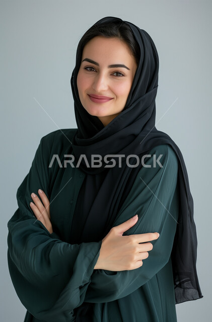 أزياء محجبات عصرية وأنيقة، الاهتمام بالمظهر الخارجي، النظر الى الكاميرا بإيماءات البهجة والسرور، مفهوم الأنوثة والنعومة، الملامح العربية القوية، بورتريه لشابةعربية خليجية سعودية ترتدي العباية والحجاب تقف بأيدي متشابكة، خلفية رمادية