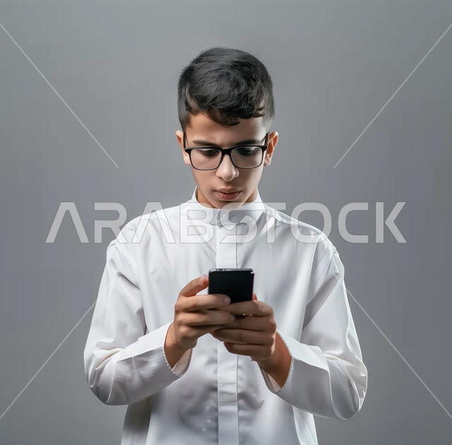 إيماءات الاندماج والتركيز، الدردشة مع الأهل والأصدقاء، تطور التكنولوجيا والأجهزة التقنية، التسوق الالكتروني وتصفح برامج وتطبيقات الجوال، بورتريه لرجل عربي سعودي خليجي يرتدي الثوب الأبيض التقليدي يحمل الهاتف المحمول، خلفية رمادية
