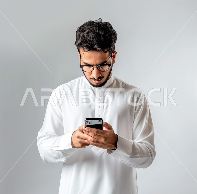 الدردشة مع الأهل والأصدقاء، تطور التكنولوجيا والأجهزة التقنية، التسوق الالكتروني وتصفح برامج وتطبيقات الجوال، بورتريه لرجل عربي سعودي خليجي يرتدي الثوب الأبيض التقليدي يحمل الهاتف المحمول، إيماءات الاندماج والتركيز، خلفية رمادية