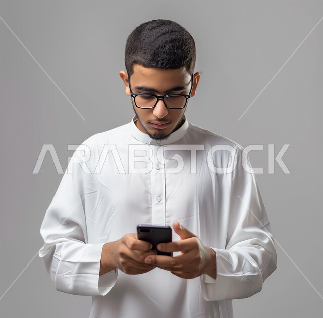 Development of technology and technical devices, online shopping and browsing mobile programs and applications, portrait of a Saudi Arabian Gulf man wearing the traditional white thobe holding a mobile phone, chatting with family and friends, gestures of integration and focus, gray background