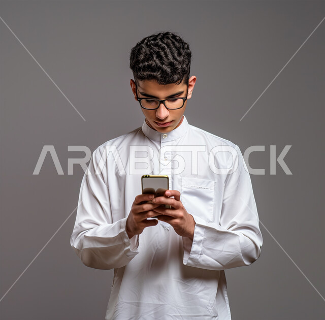 التسوق الالكتروني وتصفح برامج وتطبيقات الجوال، بورتريه لرجل عربي سعودي خليجي يرتدي الثوب الأبيض التقليدي يحمل الهاتف المحمول، تطور التكنولوجيا والأجهزة التقنية، الدردشة مع الأهل والأصدقاء، إيماءات الاندماج والتركيز، خلفية رمادية