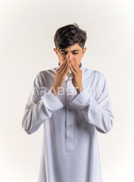 بورتريه شاب مراهق خليجي سعودي مريض يعطس داخل المناديل الورقيه بإيماءات وجه ويدين مختلفة ، يرتدي ثوب أبيض , خلفية بيضاء