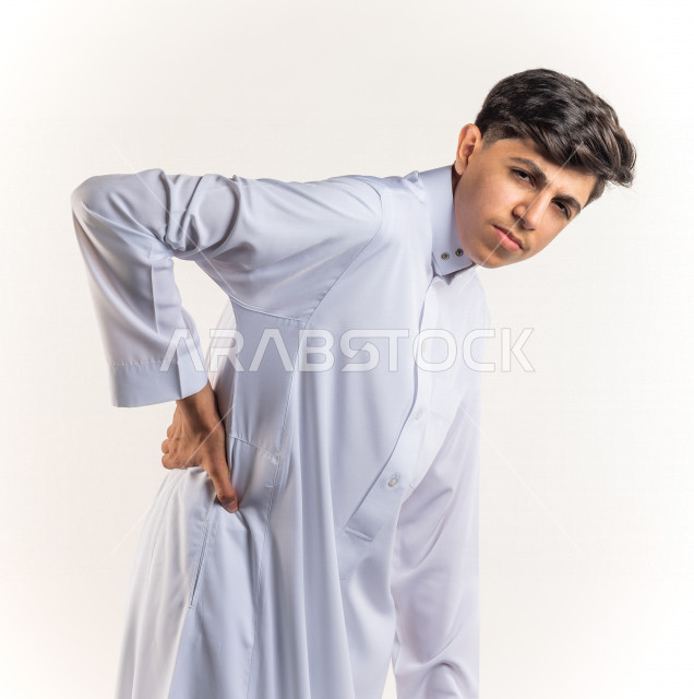 بورتريه شاب مراهق خليجي سعودي يضع يده على ظهره يشعر بحالة من الوجع والألم بإيماءات وجه ويدين مختلفة , يرتدي ثوب أبيض , خلفية بيضاء