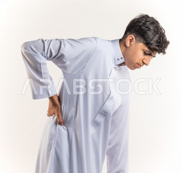 بورتريه شاب مراهق خليجي سعودي يضع يده على ظهره يشعر بحالة من الوجع والألم بإيماءات وجه ويدين مختلفة , يرتدي ثوب أبيض , خلفية بيضاء