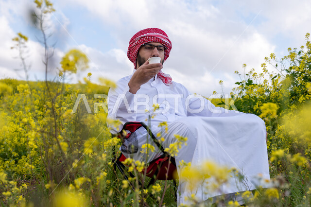 Hiking in the farms of Saudi Arabia, enjoying the natural scenery in the spring, breathing fresh air during the day, a young Saudi Arabian Gulf man wearing a traditional shemagh and thobe sitting on a chair holding a cup of coffee in his hand, gestures and expressions of pleasure and happiness, nature background