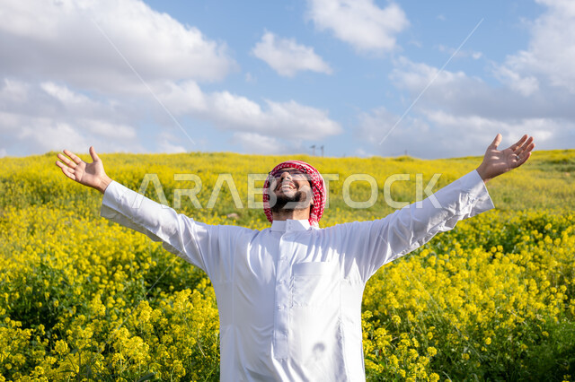 استنشاق الهواء الطلق في النهار، صورة مقربة لشاب عربي خليجي سعودي يرتدي الشماغ والثوب التقليدي يرفع يديه لأعلى، ايماءات وتعابير السرور والسعادة، التنزه في مزارع المملكة العربية السعودية، الاستمتاع بالمناظر الطبيعية في فصل الربيع، خلفية الطبيعة