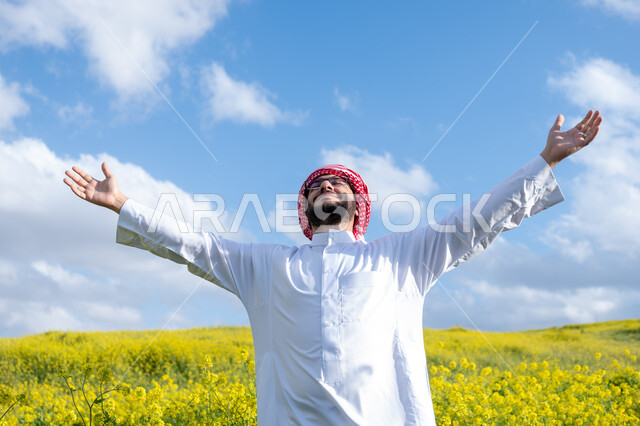 استنشاق الهواء الطلق في النهار، صورة مقربة لشاب عربي خليجي سعودي يرتدي الشماغ والثوب التقليدي يرفع يديه لأعلى، ايماءات وتعابير السرور والسعادة، التنزه في مزارع المملكة العربية السعودية، الاستمتاع بالمناظر الطبيعية في فصل الربيع، خلفية الطبيعة