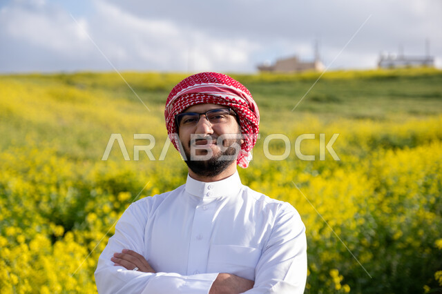 الوقوف بأيدي متقاطعة وايماءات السرور والسعادة، التنزه في مزارع المملكة العربية السعودية، الاستمتاع بالمناظر الطبيعية في فصل الربيع،  صورة مقربة لشاب عربي خليجي سعودي يرتدي الشماغ والثوب التقليدي يقوم باستنشاق الهواء الطلق في النهار، خلفية الطبيعة