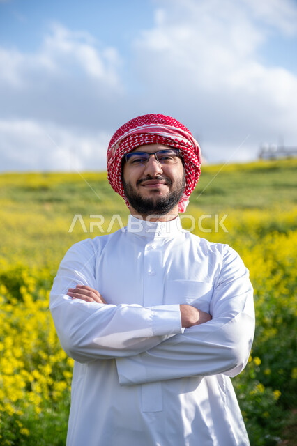 Standing with hands crossed and gestures of joy and happiness, walking in the farms of Saudi Arabia, enjoying the natural scenery in the spring, close-up of a young Saudi Arabian Gulf man wearing a traditional shemagh and thobe breathing fresh air during the day, nature background