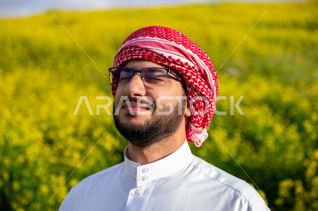 Gestures and expressions of pleasure and happiness, walking in the farms of Saudi Arabia, enjoying the natural scenery in the spring, close-up of a young Saudi Arabian Gulf man wearing a traditional shemagh and thobe breathing fresh air during the day, nature background