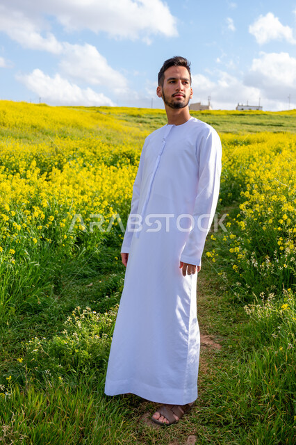 الوقوف باستقامة وبتعابير وايماءات السرور والسعادة، التنزه في مزارع الإمارات العربية المتحدة، الاستمتاع بالمناظر الطبيعية في فصل الربيع، شاب عربي خليجي إماراتي مبتسم يرتدي الكندورة يقوم باستنشاق الهواء الطلق في النهار، صورة الجسم بالكامل، خلفية الطبيعة