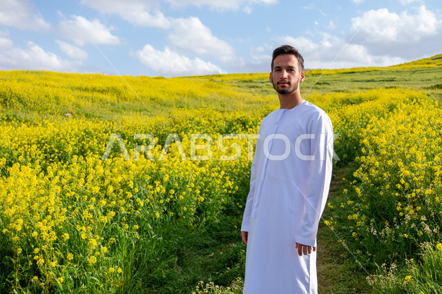 الوقوف باستقامة وبتعابير وايماءات السرور والسعادة، التنزه في مزارع الإمارات العربية المتحدة، الاستمتاع بالمناظر الطبيعية في فصل الربيع، صورة مقربة لشاب عربي خليجي إماراتي مبتسم يرتدي الكندورة يقوم باستنشاق الهواء الطلق في النهار، خلفية الطبيعة