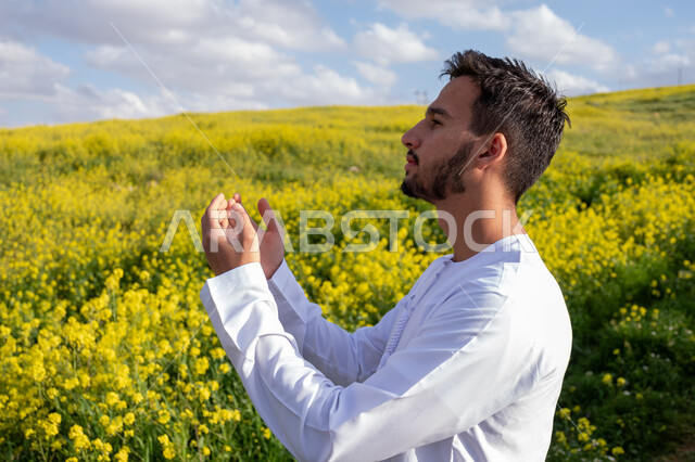 الدعاء والتضرع بتعابير الخشوع والتذلل، التنزه في مزارع الإمارات العربية المتحدة، الاستمتاع بالمناظر الطبيعية في فصل الربيع، صورة مقربة لشاب عربي خليجي إماراتي يرتدي الكندورة يرفع يديه لأعلى ويدعو الله، استنشاق الهواء الطلق في النهار، خلفية الطبيعة