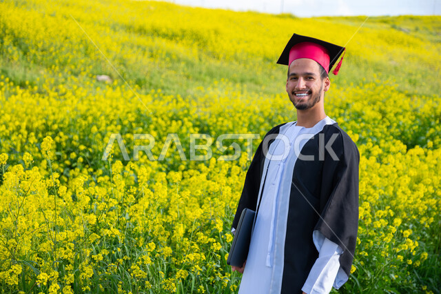 تحقيق حلم النجاح من الجامعة، المنتزهات الطبيعية في الامارات، الاحتفال بيوم التخرج بإيماءات السعادة والحماس، صورة مقربة لشاب عربي خليجي إماراتي مبتسم يرتدي الكندورة وزي التخرج مع القبعة في الخارج، الاستمتاع بالمناظر الطبيعية في فصل الربيع، خلفية الطبيعة