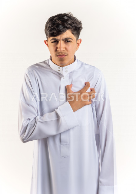 بورتريه شاب مراهق خليجي سعودي يضع يده على صدره يشعر بحالة من الوخز والألم بقلبه , خلفية بيضاء
