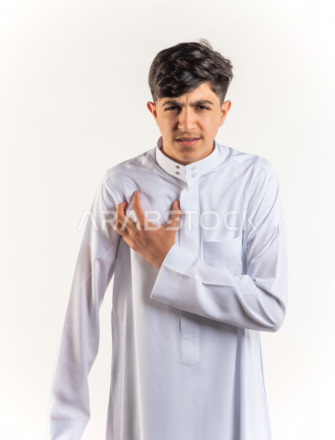 بورتريه شاب مراهق خليجي سعودي يضع يده على صدره يشعر بحالة من الوخز والألم بقلبه , خلفية بيضاء