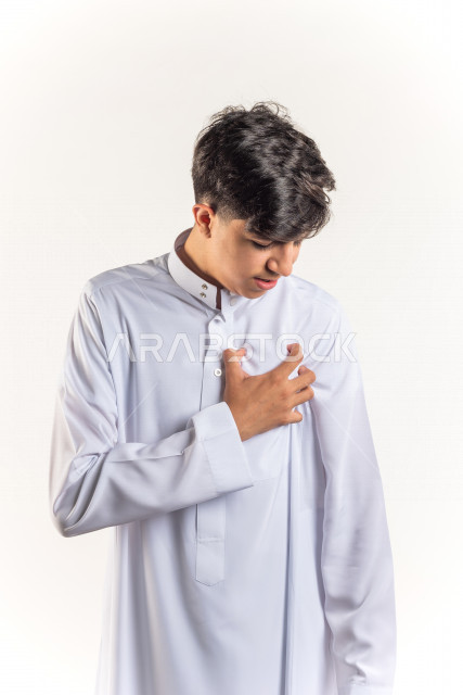 بورتريه شاب مراهق خليجي سعودي يضع يده على صدره يشعر بحالة من الوخز والألم بقلبه , خلفية بيضاء