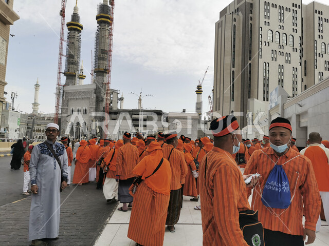 Pilgrims and Umrah performers standing in the outer courtyard of the Grand Mosque in Mecca, performing Hajj and Umrah rituals, sacred Islamic religious places and landmarks, Islamic gathering for worship and drawing closer to God, luxury hotels and towers surrounding the Grand Mosque