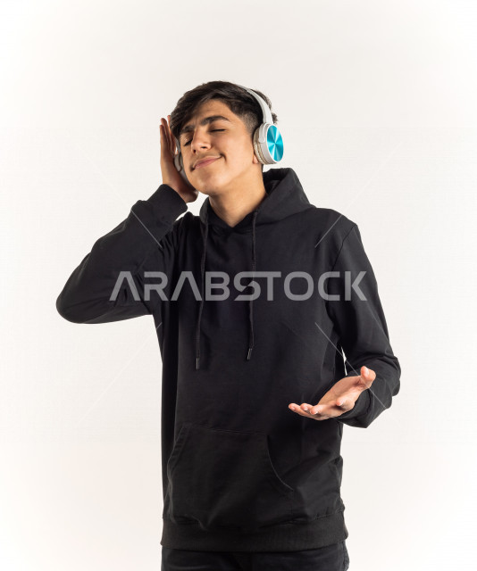 بورتريه شاب مراهق خليجي سعودي يضع سماعات على أذنيه بإيماءات وجه ويدين مختلفة تدل على الصوت العالي والتركيز , خلفية بيضاء