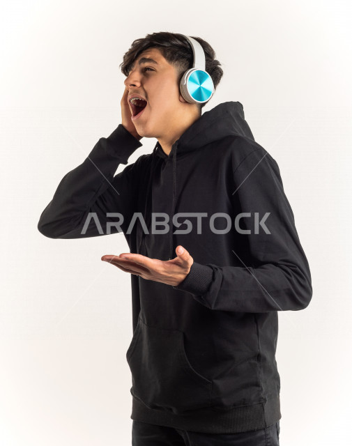 بورتريه شاب مراهق خليجي سعودي يضع سماعات على أذنيه بإيماءات وجه ويدين مختلفة تدل على الصوت العالي والتركيز , خلفية بيضاء