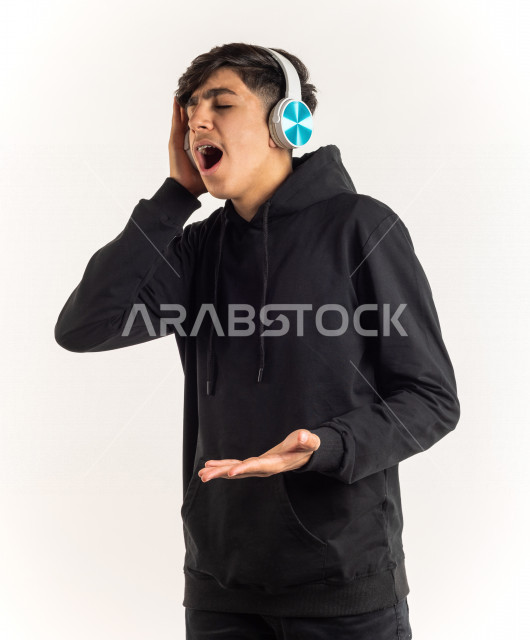 بورتريه شاب مراهق خليجي سعودي يضع سماعات على أذنيه بإيماءات وجه ويدين مختلفة تدل على الصوت العالي والتركيز , خلفية بيضاء
