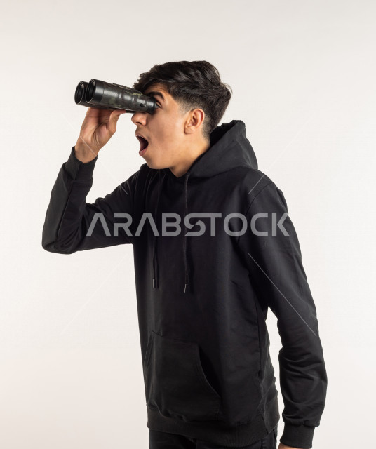 بورتريه شاب مراهق خليجي سعودي يقوم بالنظر من خلال المنظار ثنائي العينين (التلسكوب) , خلفية بيضاء
