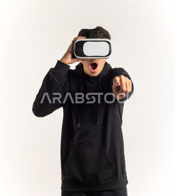 بورتريه شاب مراهق خليجي سعودي يقوم بالنظر من خلال نظارات الواقع الإفتراضي (في آر بوكس) بإيماءات وجه ويدين مختلفة , خلفية بيضاء