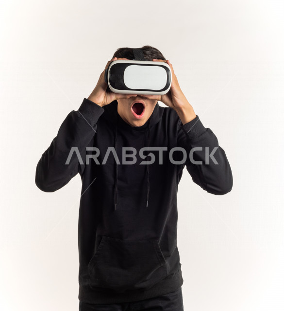 بورتريه شاب مراهق خليجي سعودي يقوم بالنظر من خلال نظارات الواقع الإفتراضي (في آر بوكس) بإيماءات وجه ويدين مختلفة , خلفية بيضاء