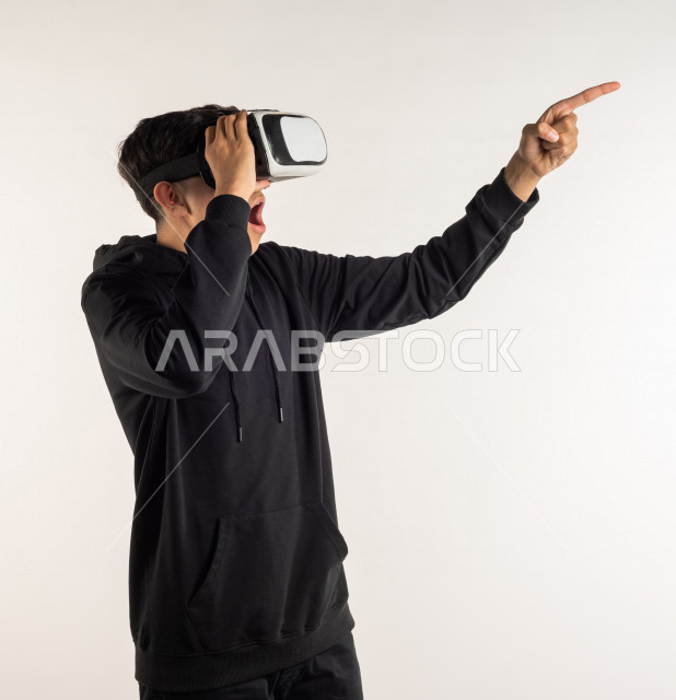 بورتريه شاب مراهق خليجي سعودي يقوم بالنظر من خلال نظارات الواقع الإفتراضي (في آر بوكس) بإيماءات وجه ويدين مختلفة , خلفية بيضاء