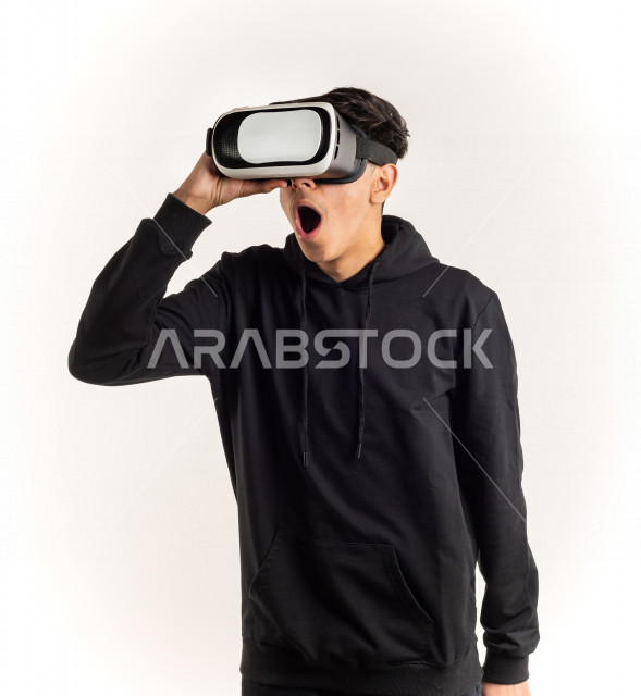 بورتريه شاب مراهق خليجي سعودي يقوم بالنظر من خلال نظارات الواقع الإفتراضي (في آر بوكس) بإيماءات وجه ويدين مختلفة , خلفية بيضاء