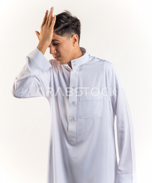 بورتريه شاب مراهق خليجي سعودي مريض يضع يده على رأسه بإيماءات وجه ويدين مختلفة تدل على الألم و الوجع ، خلفية بيضاء