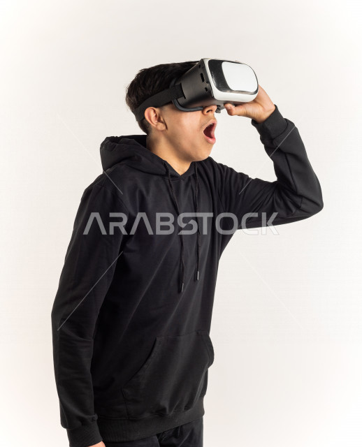 بورتريه شاب مراهق خليجي سعودي يقوم بالنظر من خلال نظارات الواقع الإفتراضي (في آر بوكس) بإيماءات وجه ويدين مختلفة , خلفية بيضاء