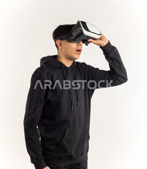بورتريه شاب مراهق خليجي سعودي يقوم بالنظر من خلال نظارات الواقع الإفتراضي (في آر بوكس) بإيماءات وجه ويدين مختلفة , خلفية بيضاء