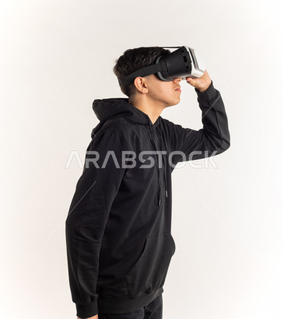 بورتريه شاب مراهق خليجي سعودي يقوم بالنظر من خلال نظارات الواقع الإفتراضي (في آر بوكس) بإيماءات وجه ويدين مختلفة , خلفية بيضاء