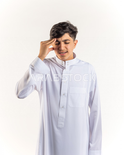 بورتريه شاب مراهق خليجي سعودي مريض يضع يده على رأسه بإيماءات وجه ويدين مختلفة تدل على الألم و الوجع ، خلفية بيضاء